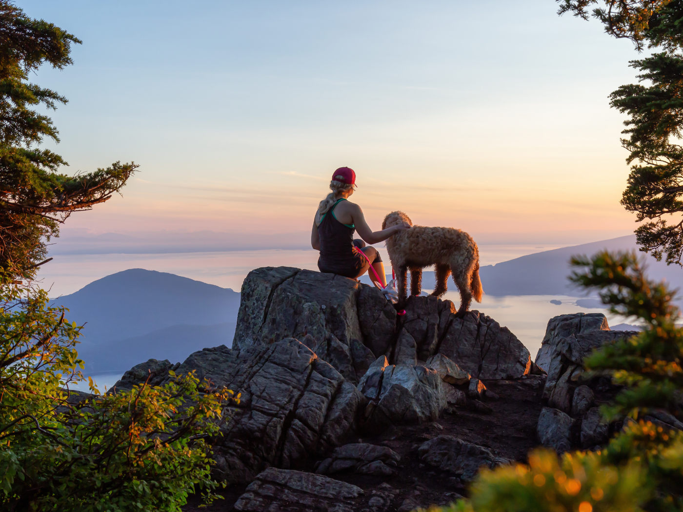 A woman and her dog sitting on a rocky peak, watching the sunset over mountains and a body of water.