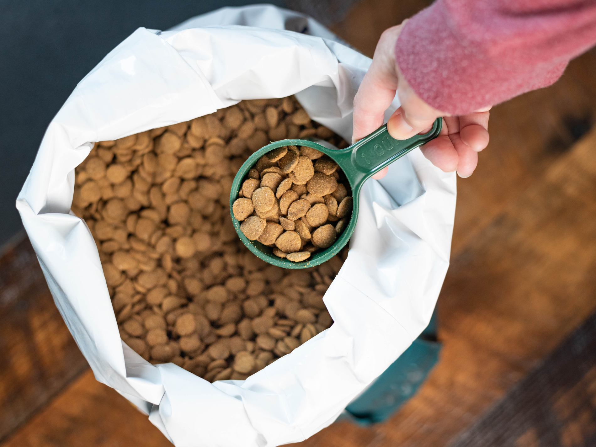 A hand holding a green measuring cup filled with dry brown kibble over an open bag of pet food.