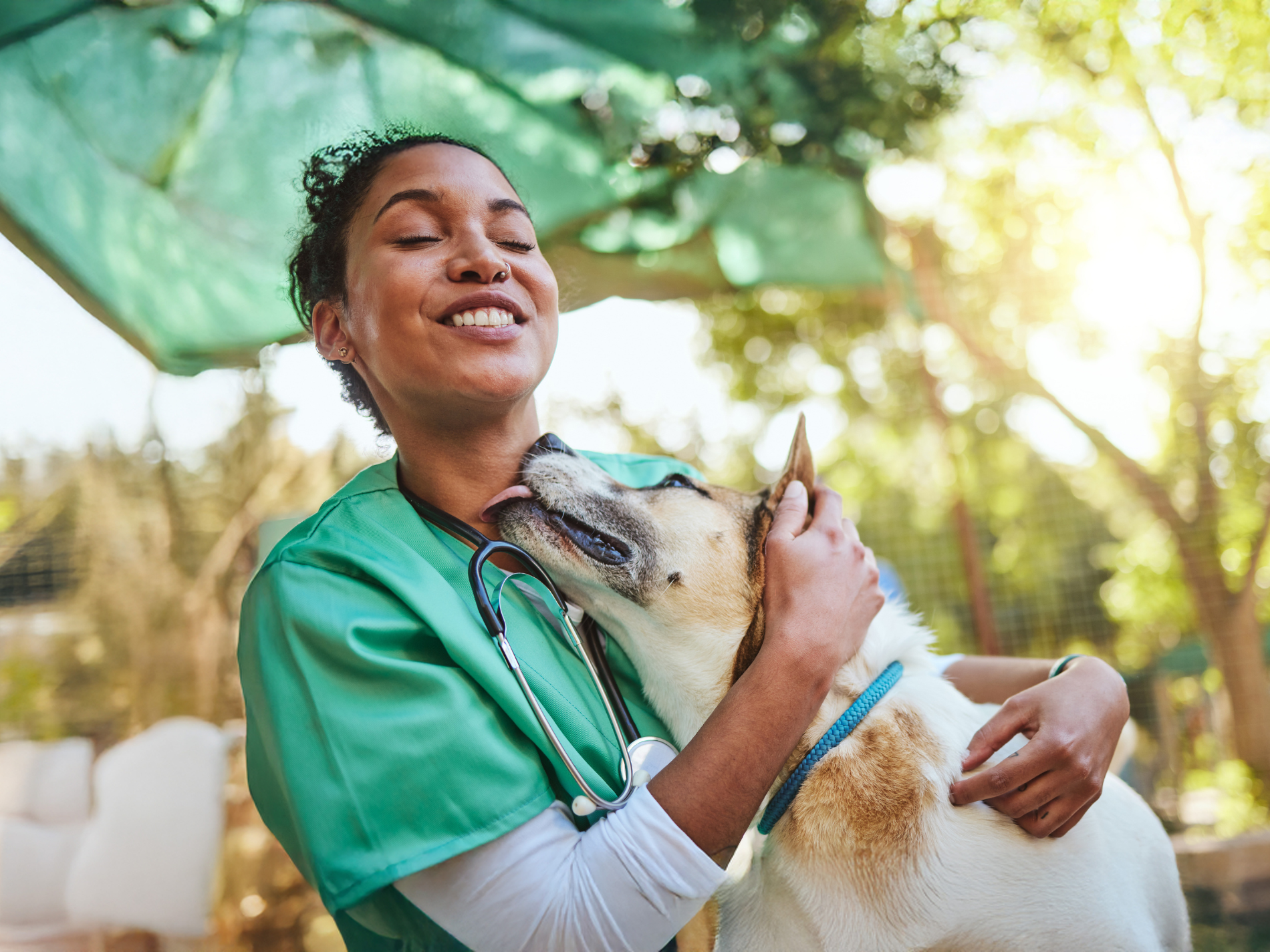 Smiling veterinarian in green scrubs being affectionately licked by a dog outdoors.