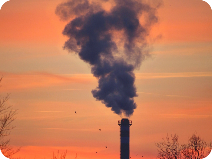 Thick dark smoke billows from a tall industrial chimney against a vibrant orange and pink sunset sky.