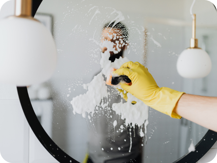 Hand in a yellow rubber glove using a sponge to clean white soap suds off a round bathroom mirror.