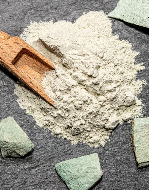 Fine pale green clinoptilolite powder with a wooden scoop on a dark textured slate surface with raw mineral chunks.